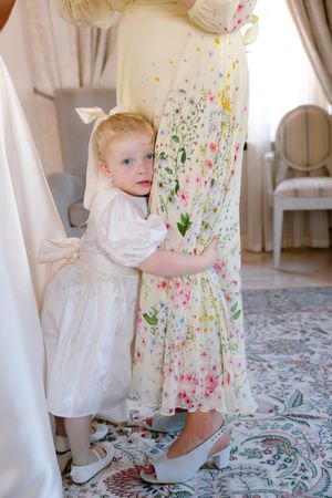 A toddler in a white dress stands next to floral patterned curtains on a patterned rug in a bright interior space.