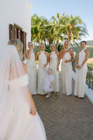 A group in white dresses and gowns pose together on a balcony with palm trees and ocean views in the background.