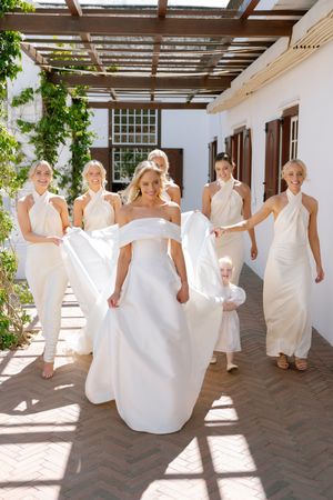 A group in flowing white dresses dances together in a sunlit courtyard with brick flooring and white walls.