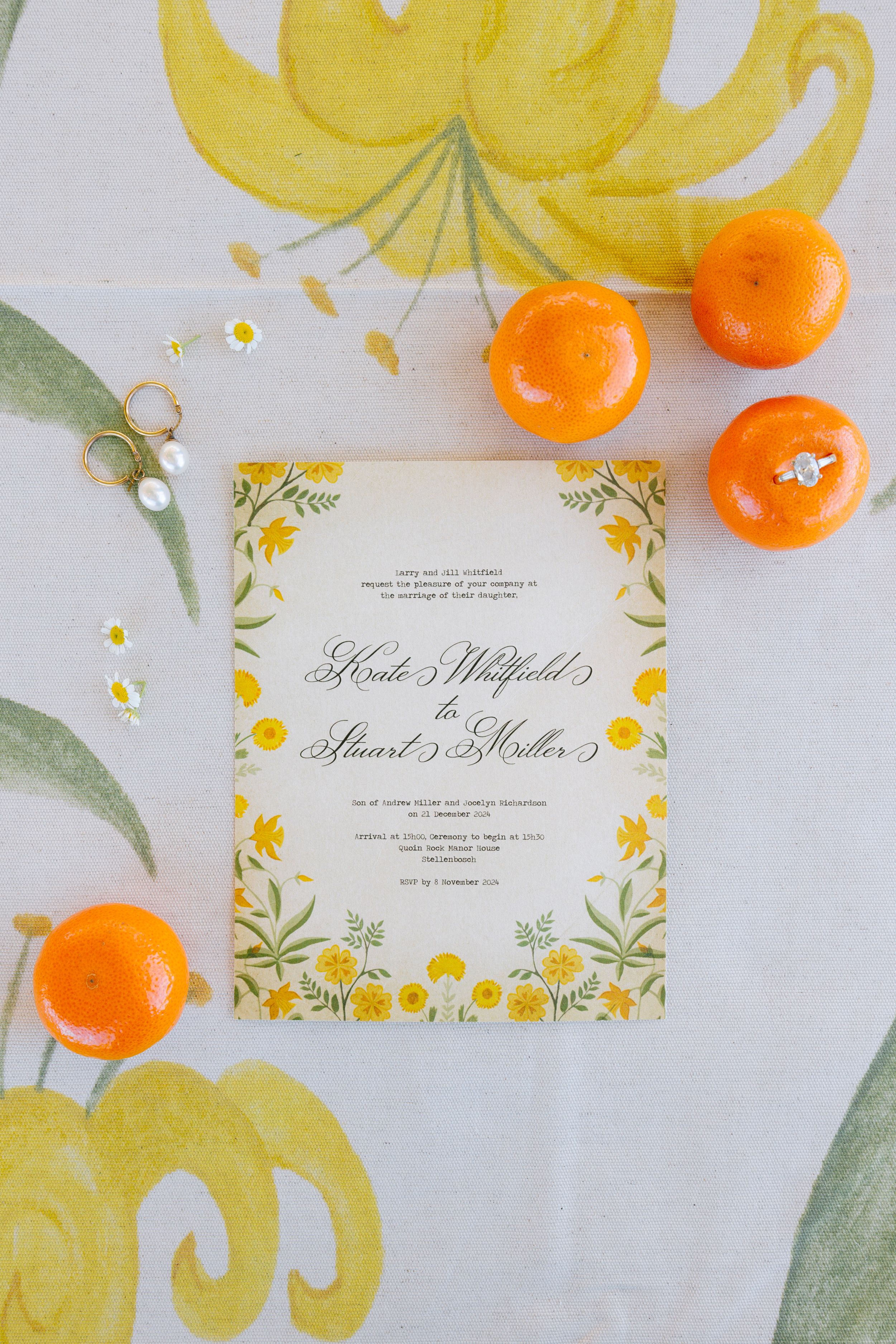 Wedding invitation cards with yellow floral designs laid out with fresh orange citrus fruits on a light gray surface.