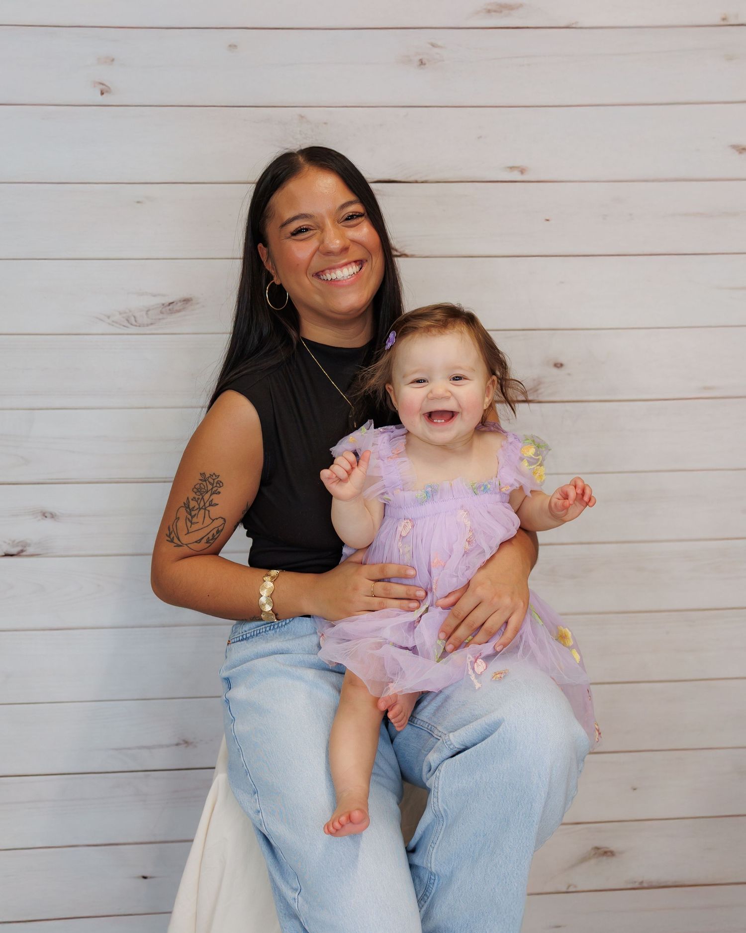 A joyful moment captured against a white wood panel backdrop showing a parent and laughing baby in a pink dress.