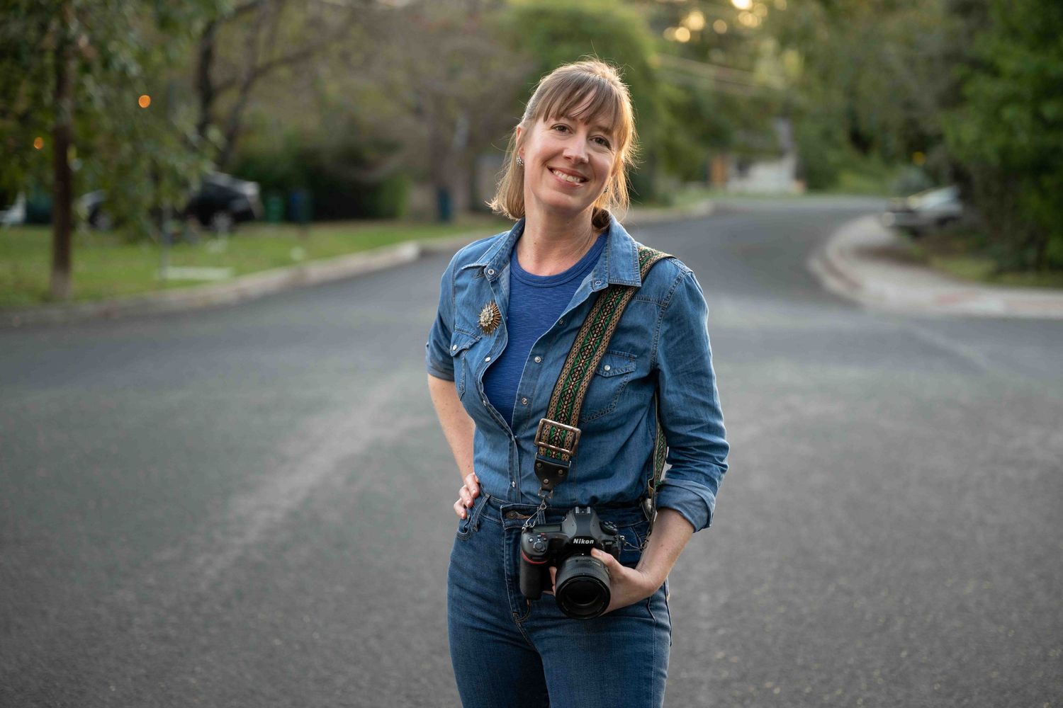 A photographer with a DSLR camera smiles while standing outdoors with people seated at tables in the background.