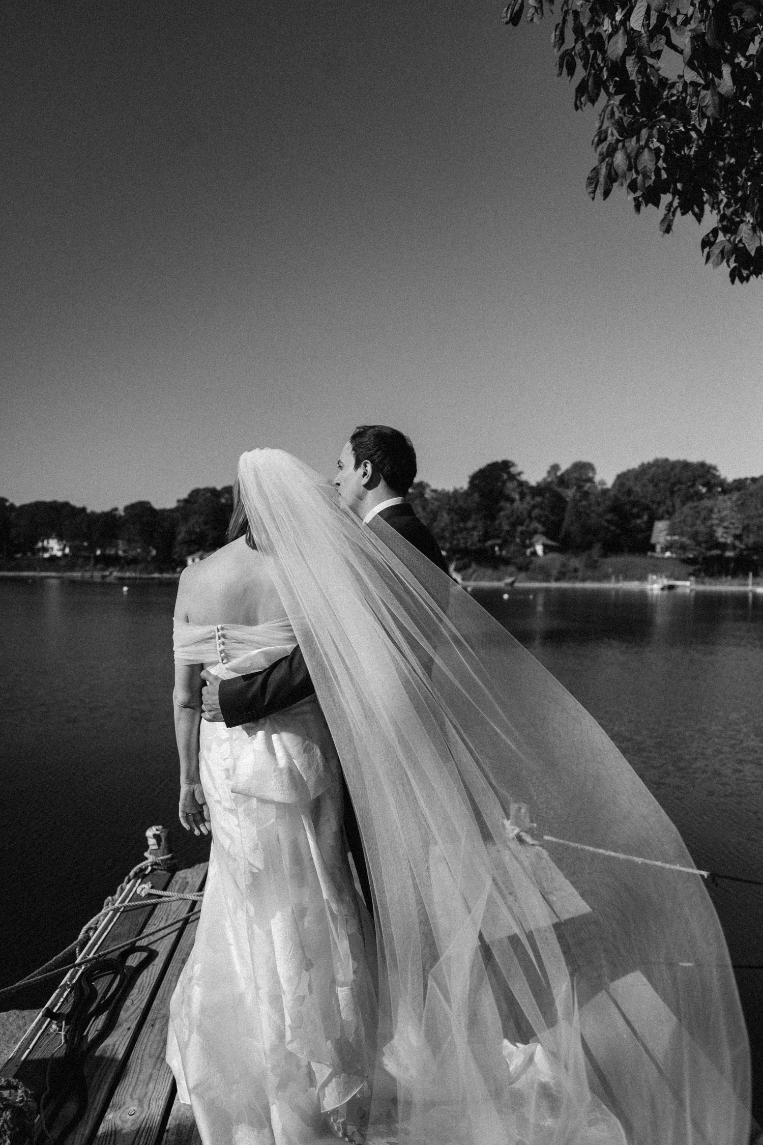 A flowing white wedding veil billows dramatically in the breeze against a lakeside landscape in black and white. By Anaise Prince