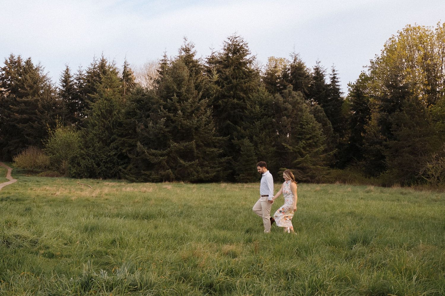 A couple and their golden retriever walk through a scenic meadow at sunset near evergreen trees.