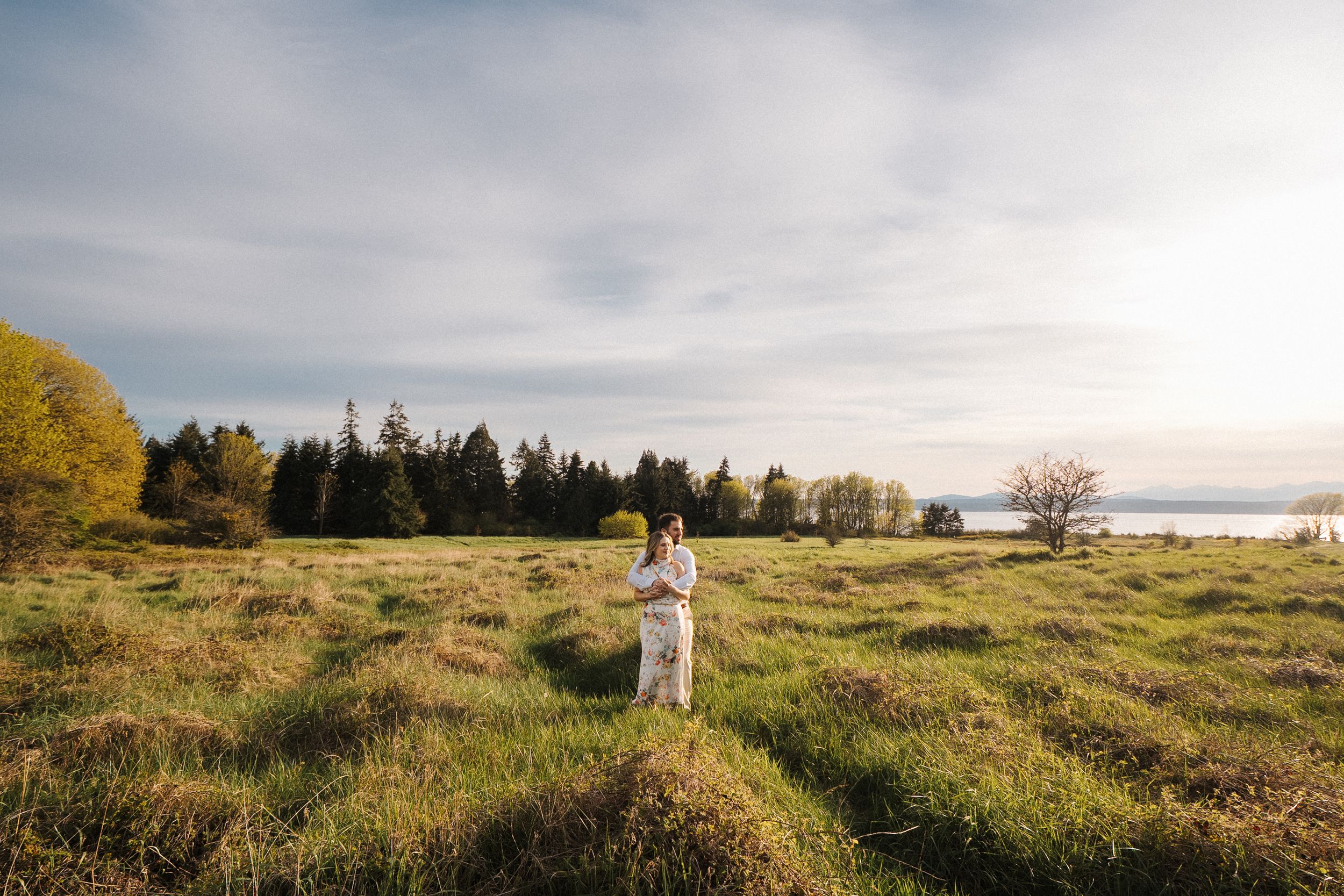 A couple stands together in a golden meadow at sunset with evergreen trees in the background.