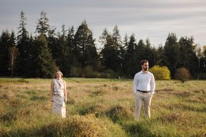 A couple stands together in a golden meadow with evergreen trees in the background during sunset.