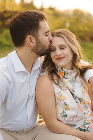 Romantic couple embraces in golden sunlight while wearing white shirt and floral dress during outdoor photo session.