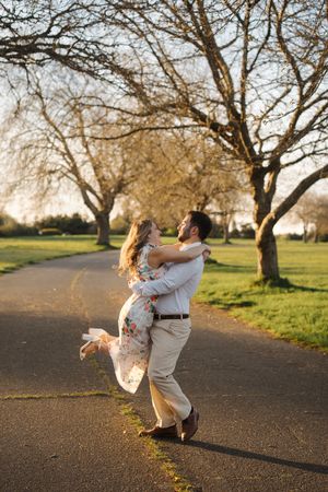A couple embraces and spins joyfully in a park at sunset among bare trees.