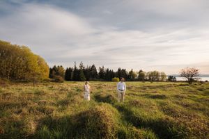 A couple walks through a golden autumn field at sunset with tall grass and trees in the background.
