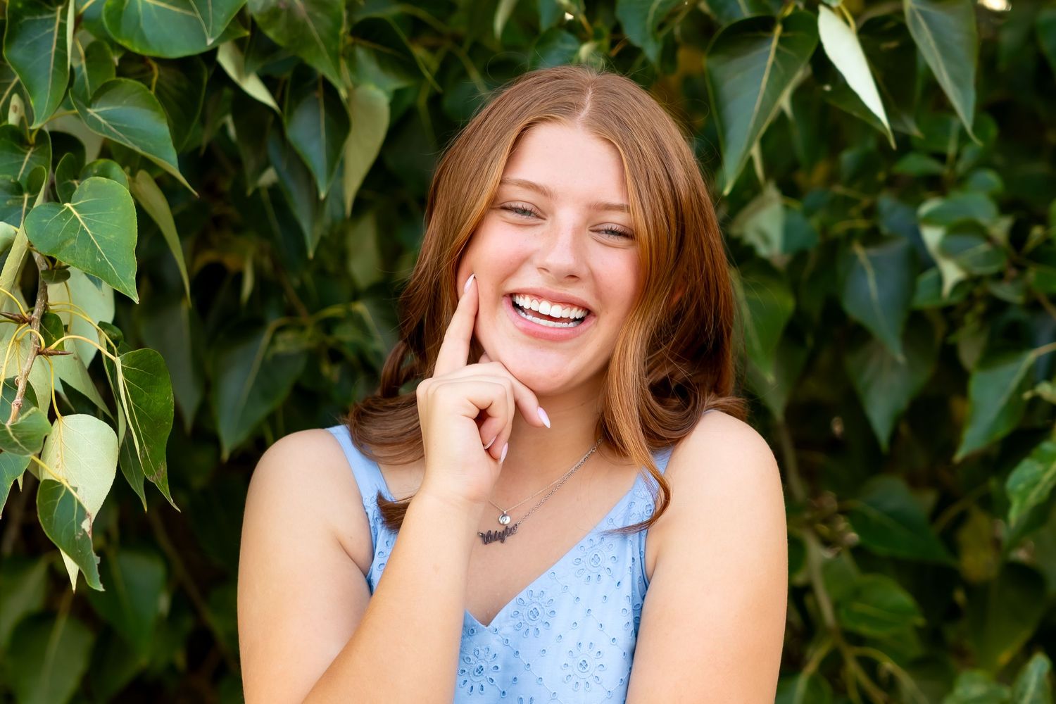 A joyful person wearing a blue polka dot top poses against a lush green leafy backdrop during a sunny outdoor photoshoot.