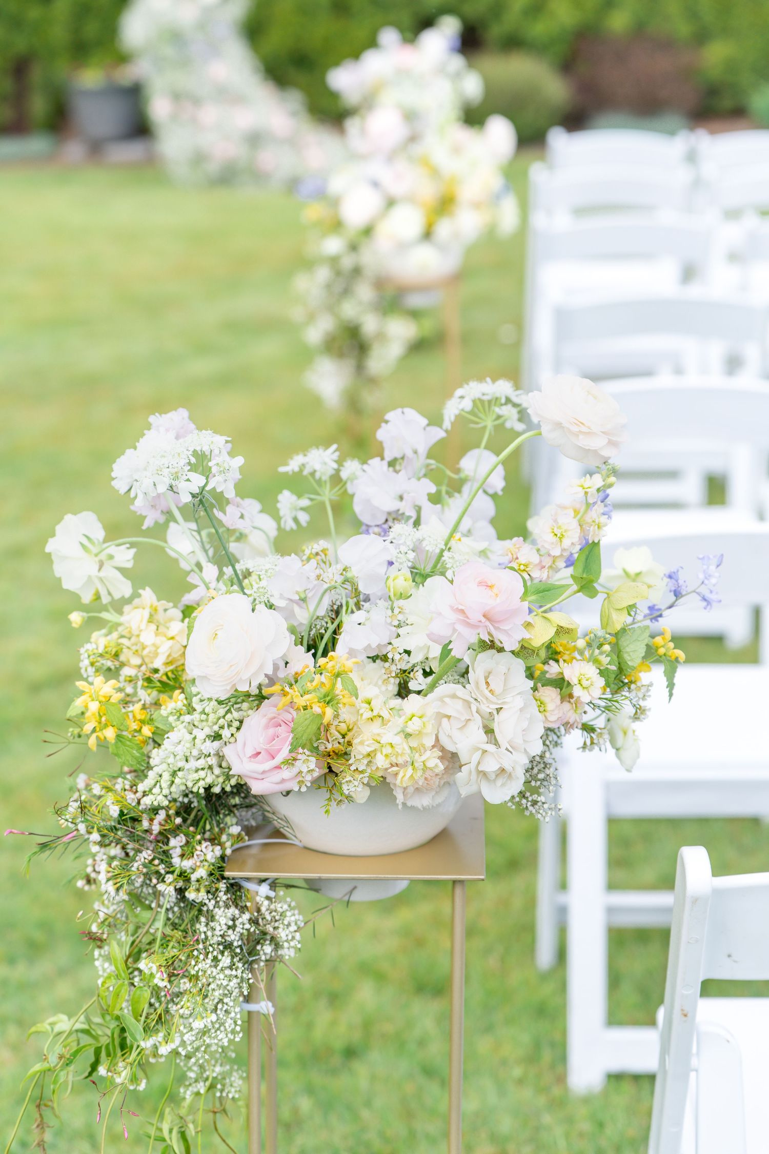 Elegant floral arrangements with pink and white flowers decorate white chairs along an outdoor wedding ceremony aisle.