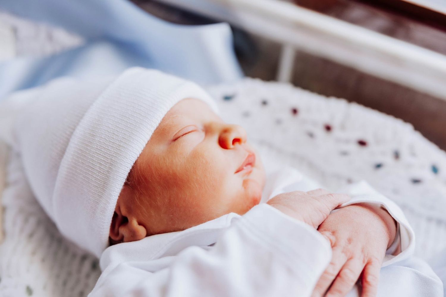 Sleeping newborn baby wearing a white hat and outfit resting in a hospital bassinet.