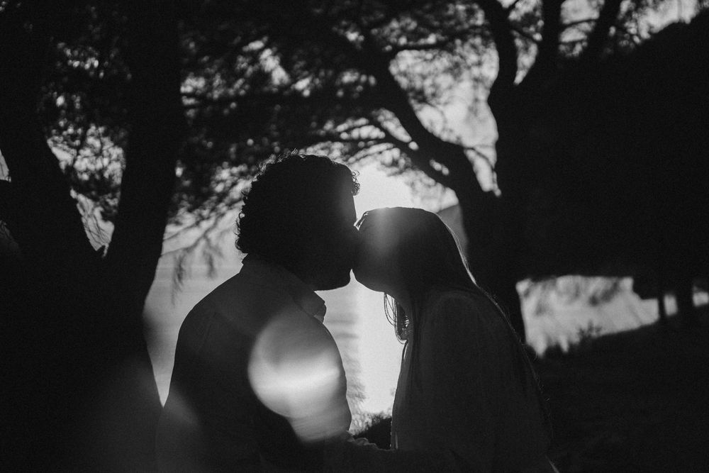 A romantic silhouette of a couple kissing at sunset with sunlight peeking through trees in black and white.