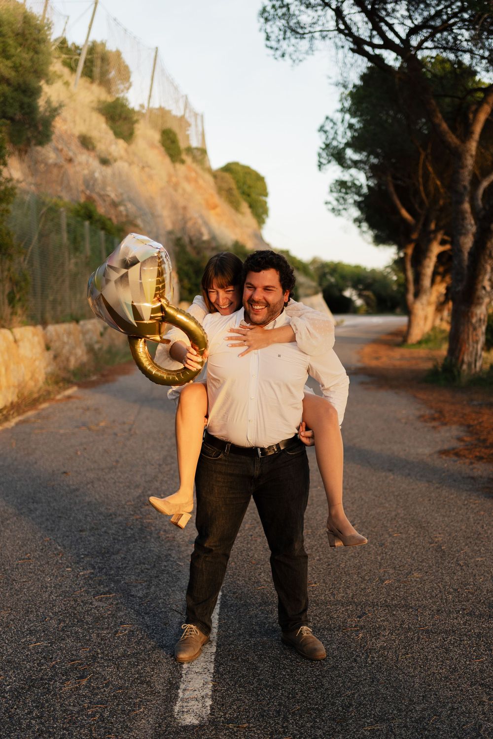 Playful couple dancing and laughing together on a tree-lined dirt road at golden hour.