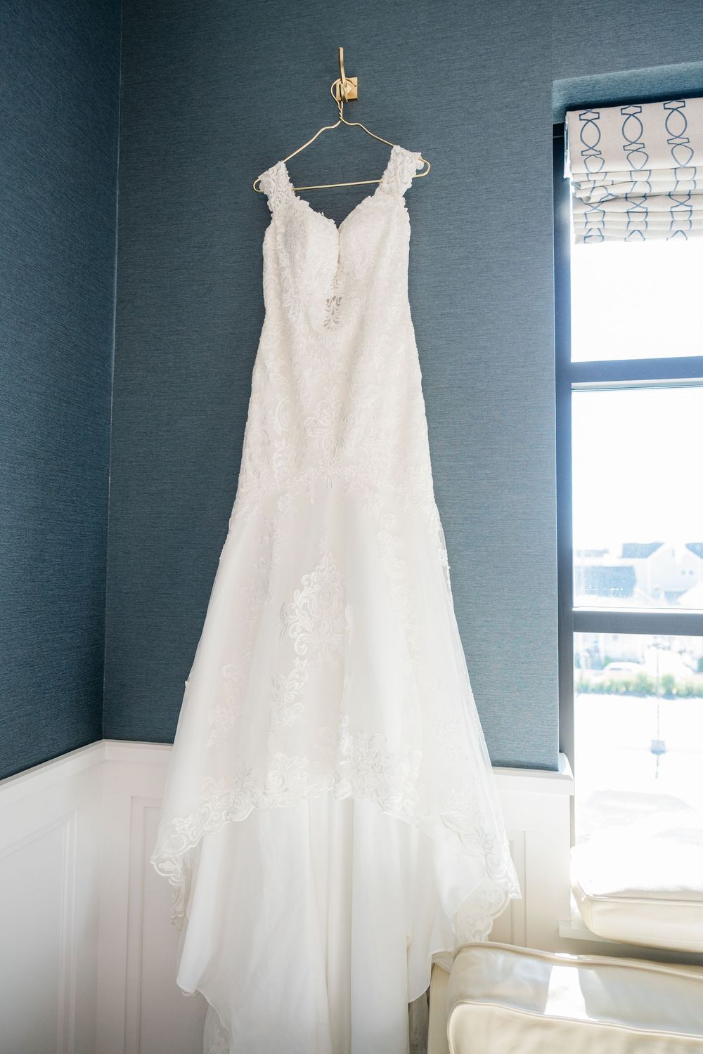 A white lace wedding dress hangs on display against a blue-gray wall near a window.