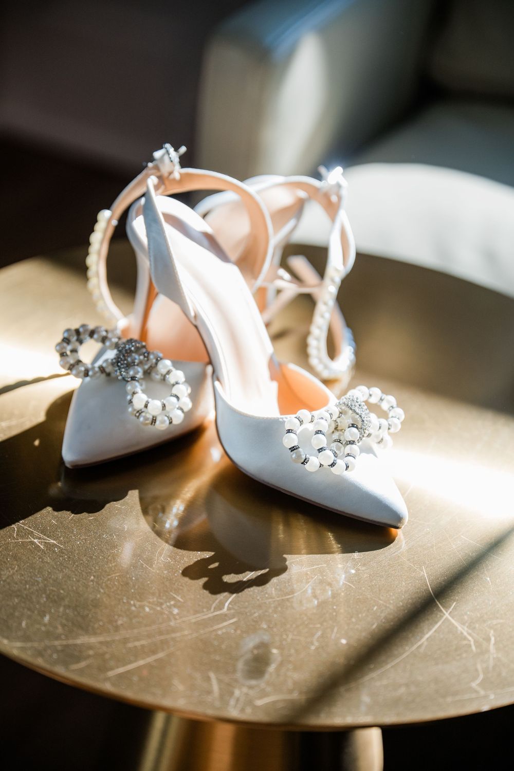 Elegant white bridal shoes with crystal embellishments displayed on a gold table in natural light.