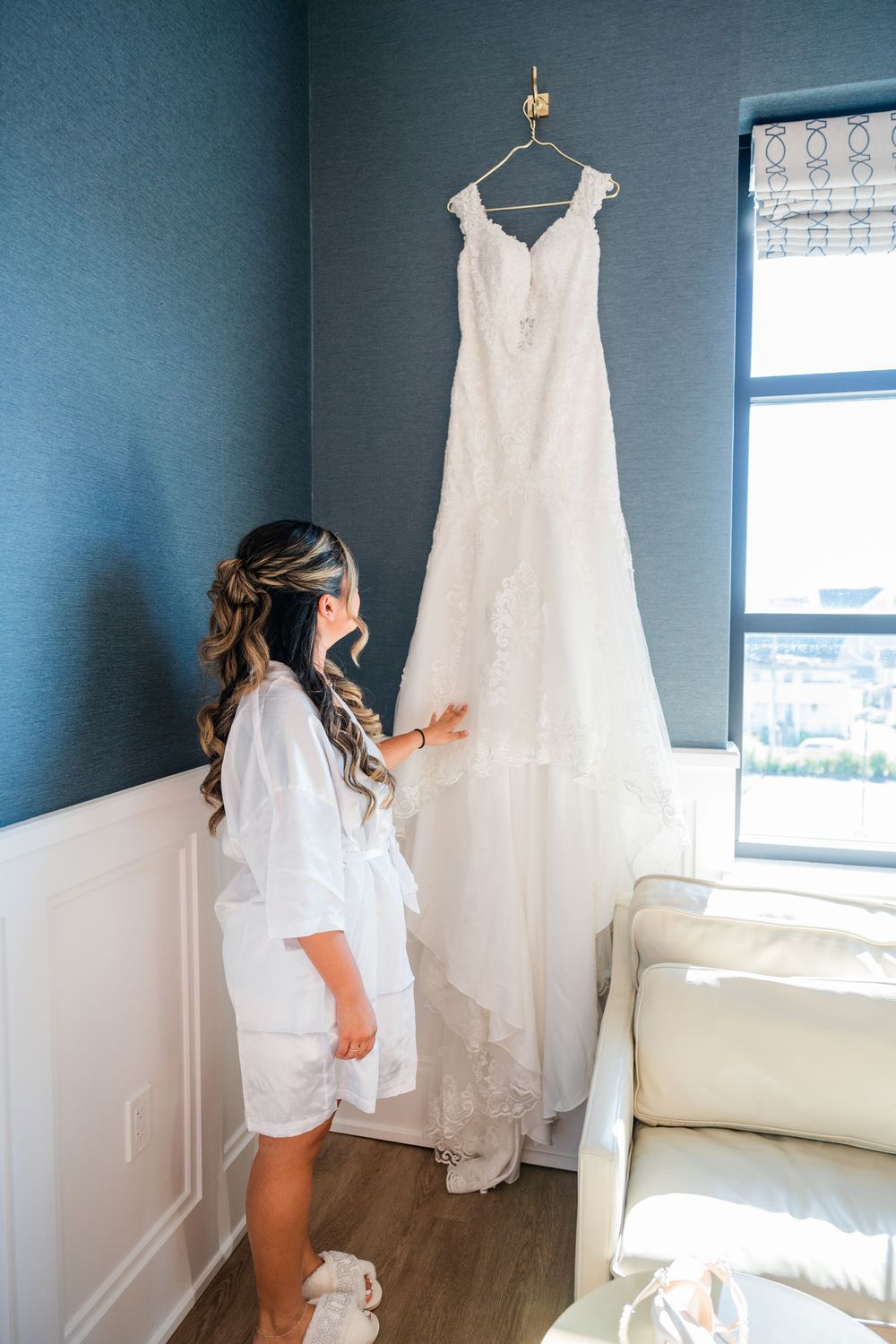 A white wedding dress hangs on display in a room with blue walls while someone in a white robe admires it.