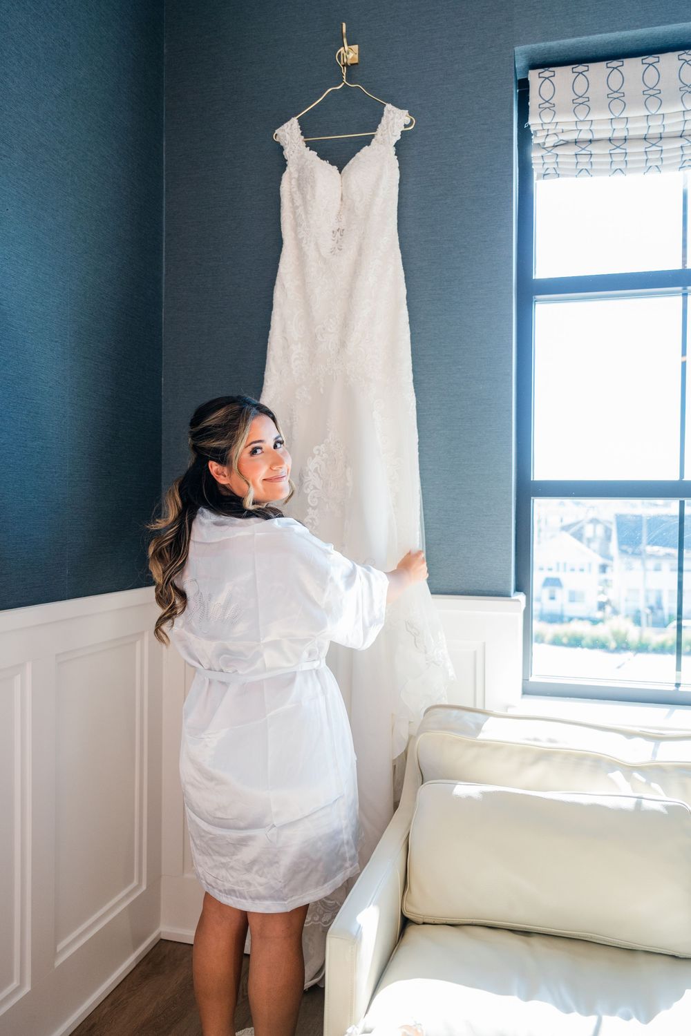 Wedding dress hanging in a blue-walled room with white wainscoting and natural light streaming through the window.