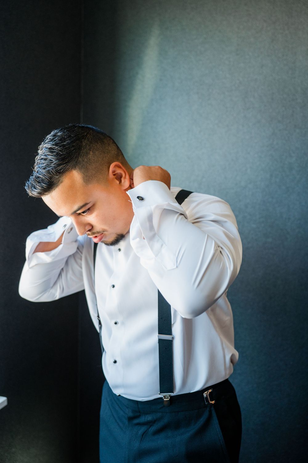 A groom in a crisp white dress shirt and suspenders adjusts his collar while getting ready.
