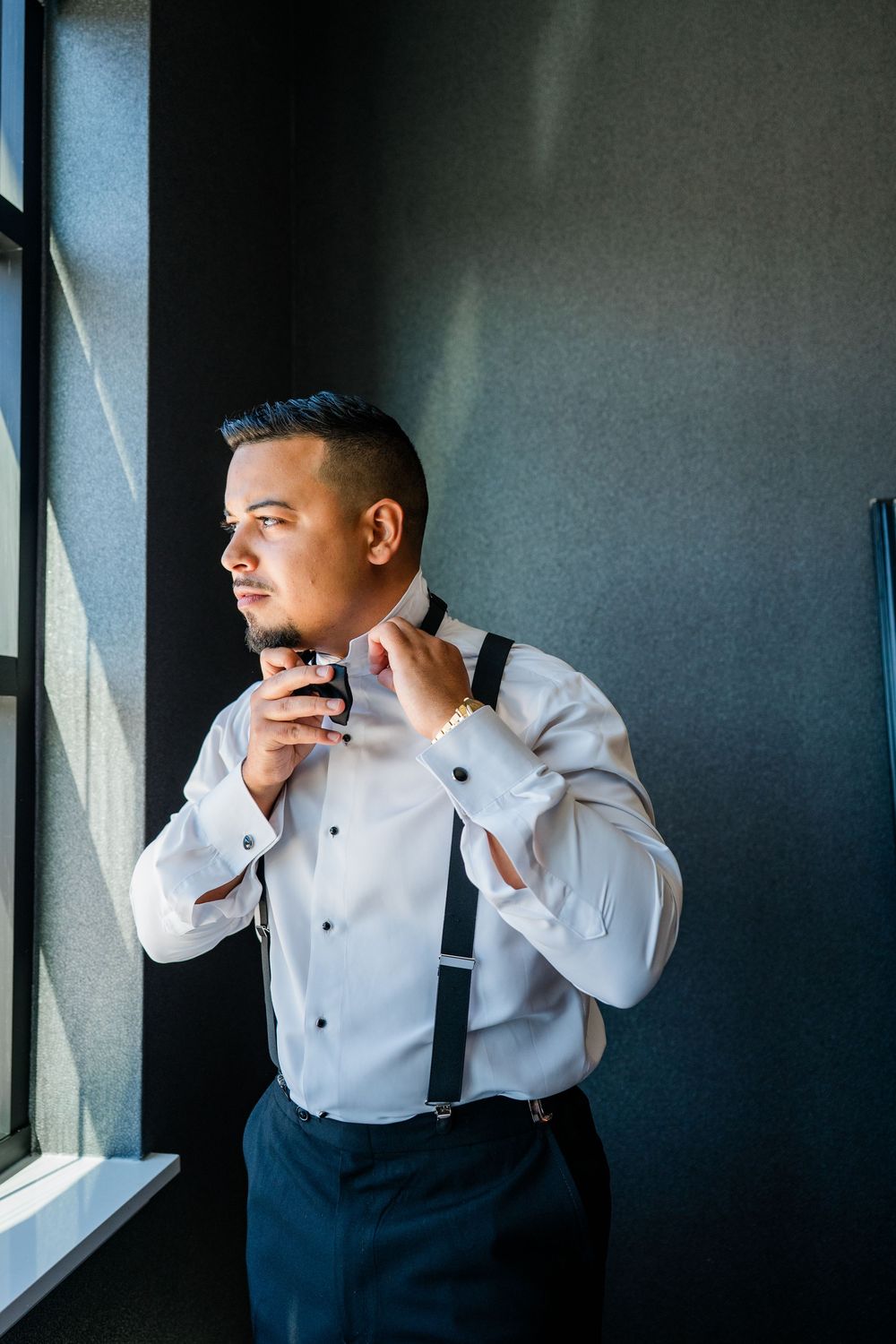 Groom in white dress shirt and suspenders gazes thoughtfully out a window in dramatic lighting.