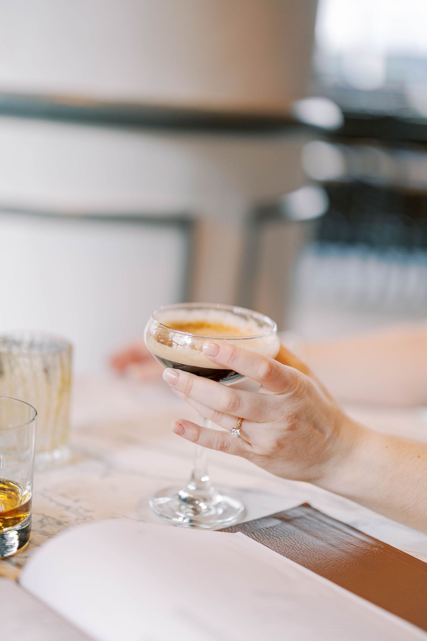 Hand holding a coupe glass filled with a light-colored cocktail at an elegant bar setting.