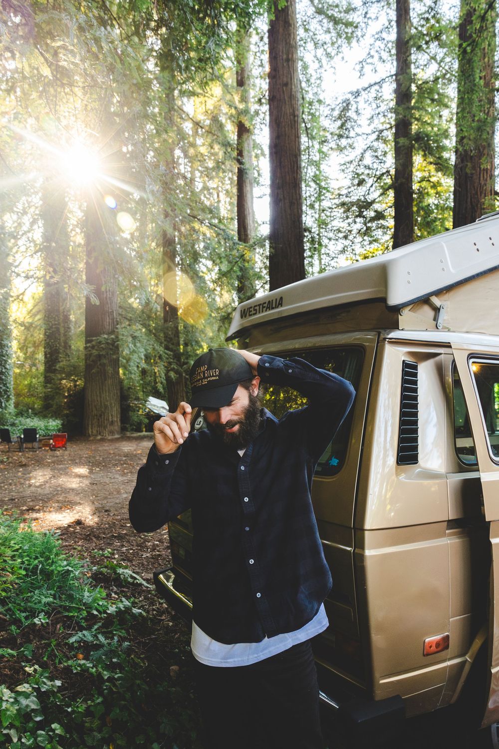 A person in a black jacket gestures with thumbs up next to a vintage van parked in a sunlit forest setting.