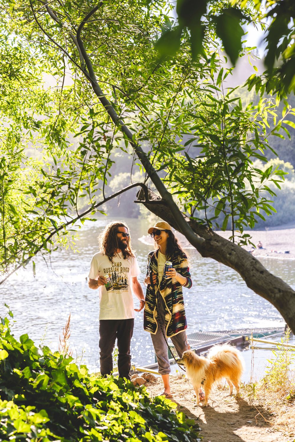 Two people and a dog enjoy a sunny afternoon by the water's edge under green tree branches.