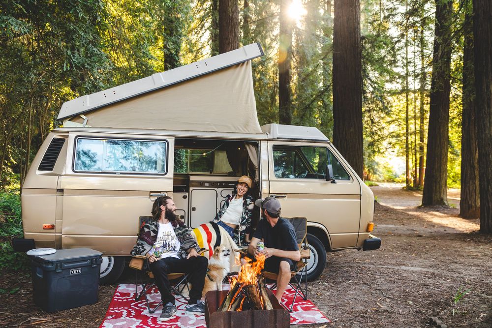 Vintage camper van parked in a sunlit forest campsite with camping chairs and blankets set up outside.