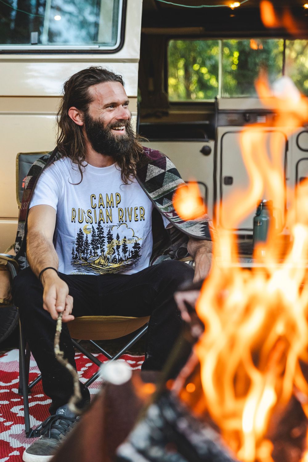 A campfire burns in the foreground while someone relaxes in a camper van wearing an outdoor-themed t-shirt.