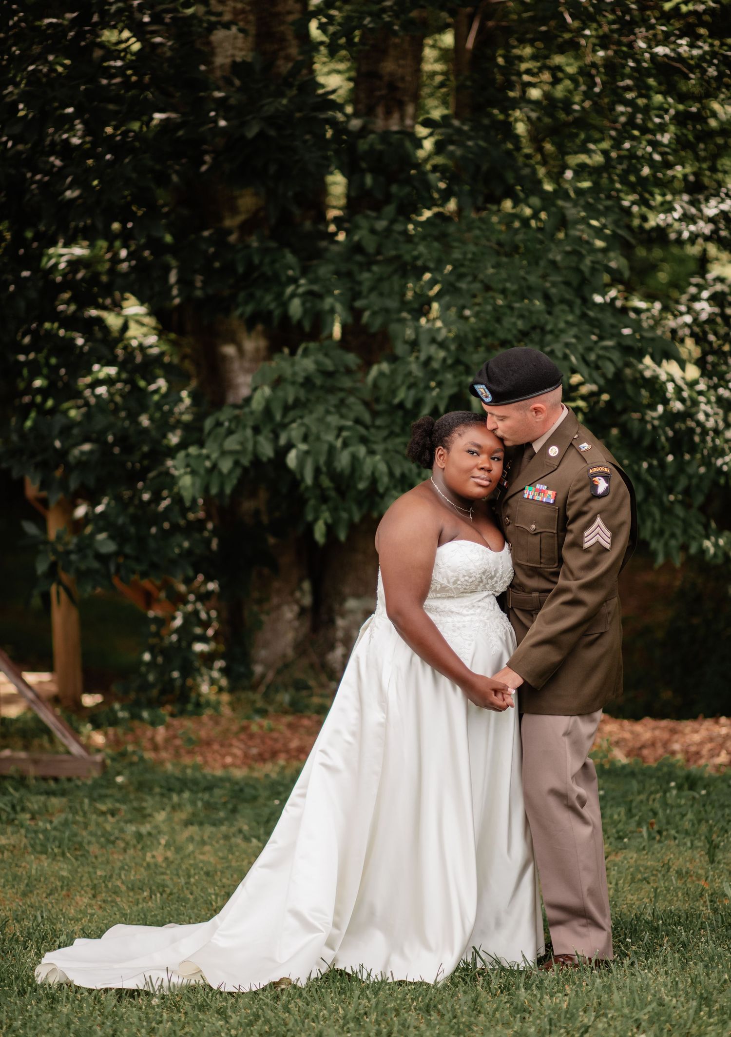 Military wedding couple shares intimate moments on grassy lawn surrounded by trees.