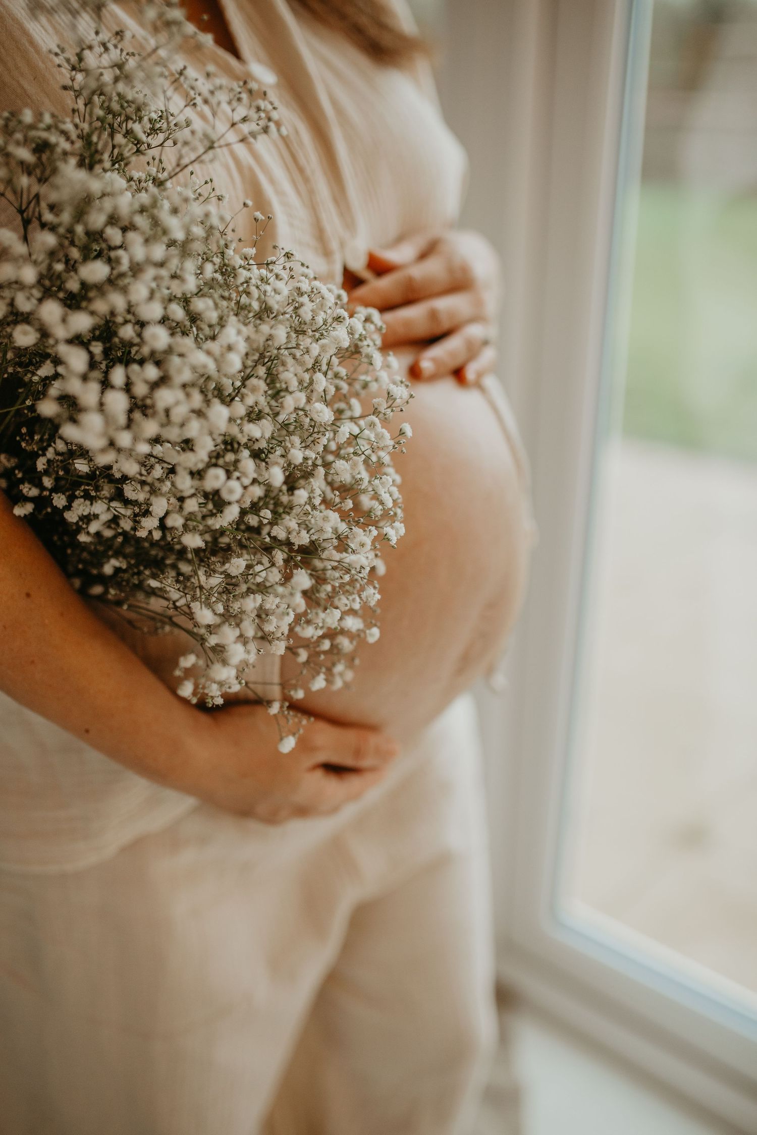 Delicate baby's breath flowers held against a soft white maternity dress near a window in natural light.