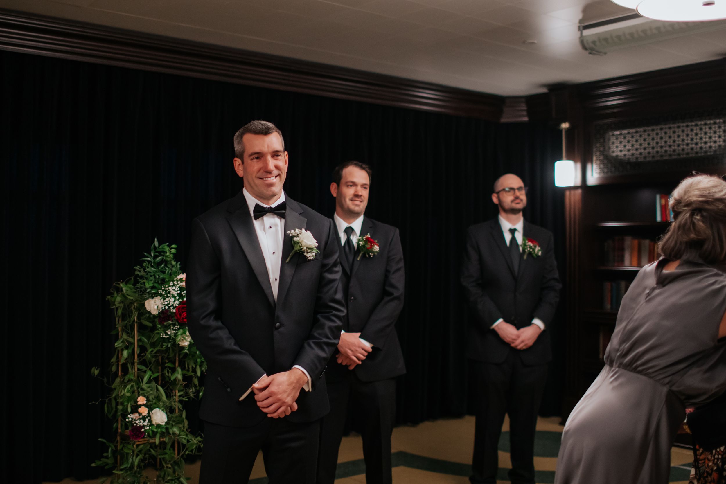 A formal wedding party in black tuxedos standing at the front of a dark-walled ceremony space.