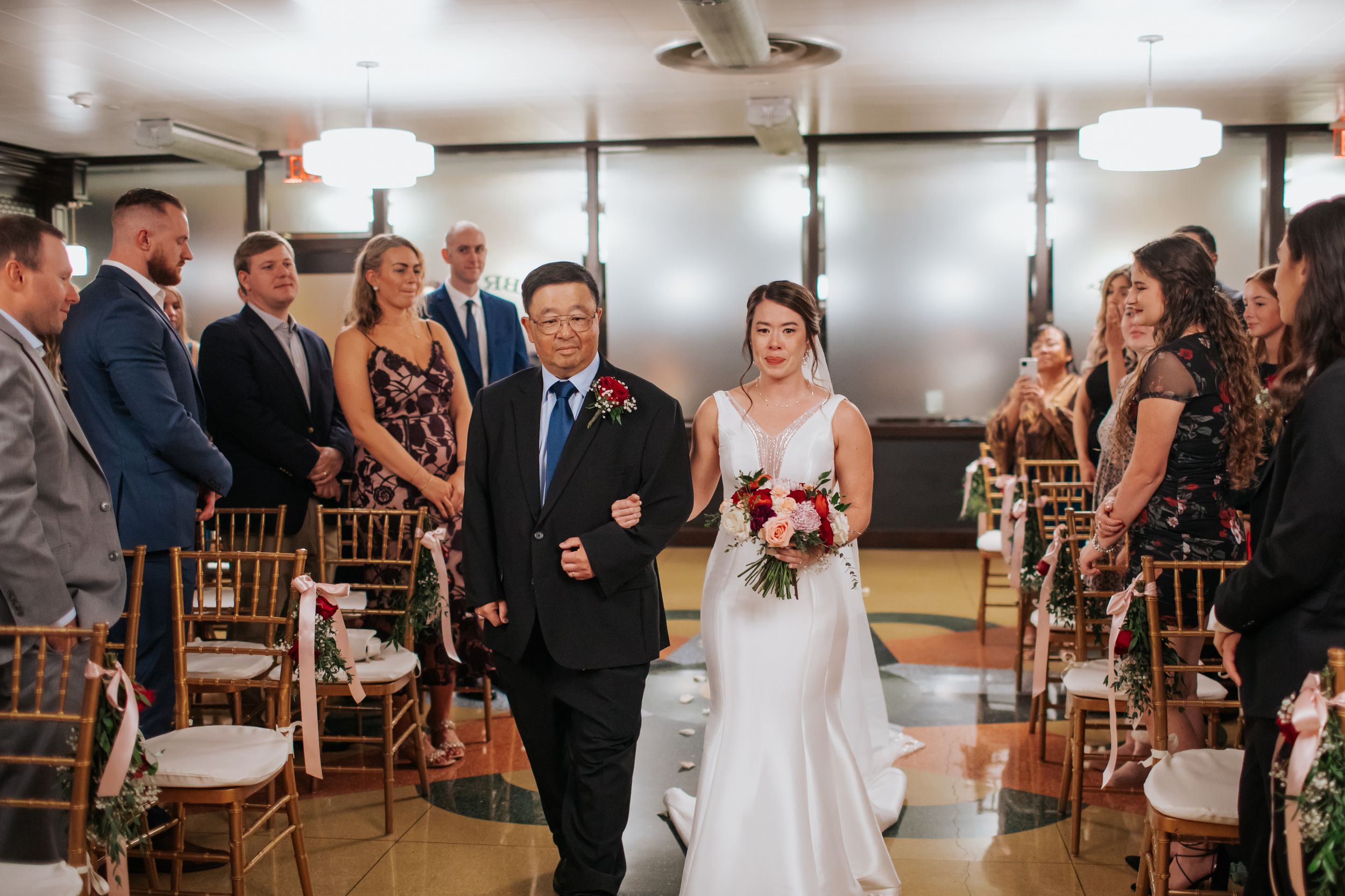 A wedding ceremony in progress inside an elegant indoor venue with gold chiavari chairs and modern lighting fixtures.