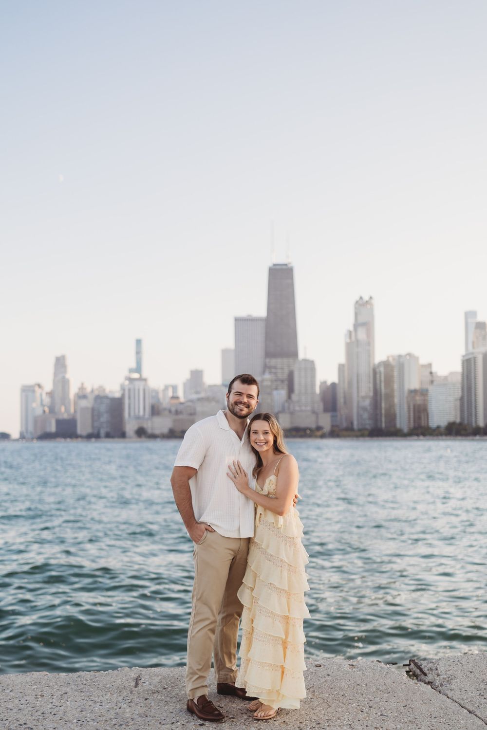 A couple poses along Lake Michigan with the iconic Chicago skyline in the background at sunset.