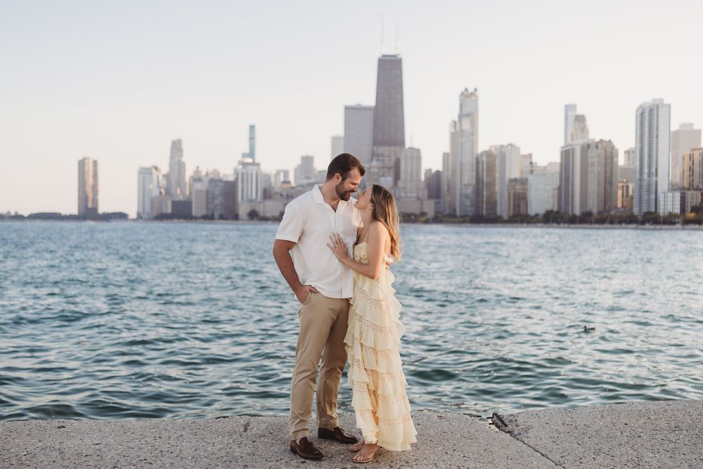 A romantic couple embraces on a beach with the Chicago skyline reflecting in Lake Michigan at sunset.