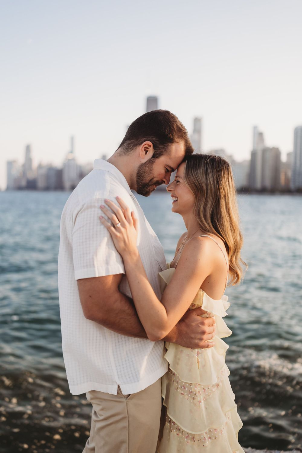 Romantic couple embraces by Lake Michigan with Chicago skyline in background during sunset photoshoot.