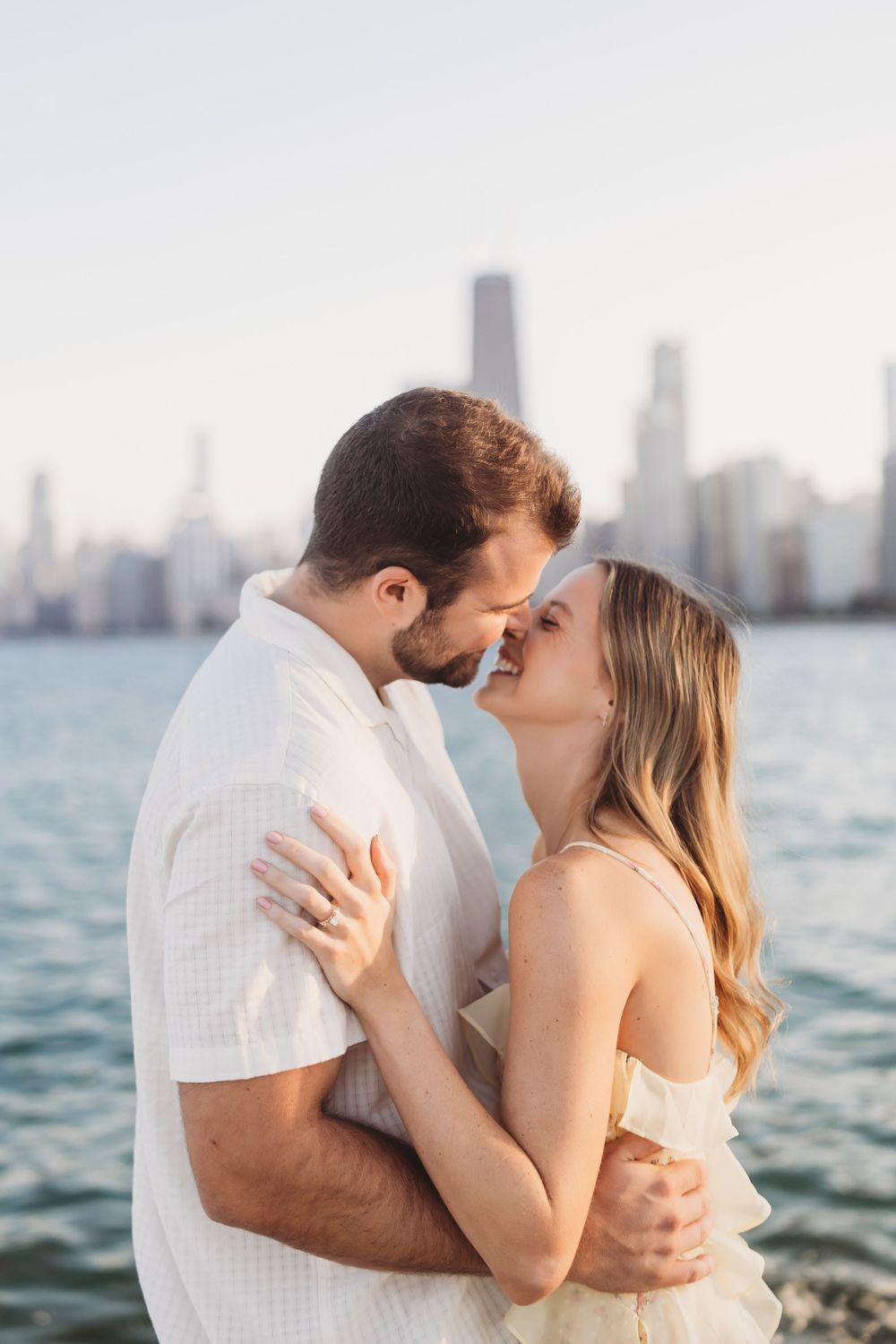 Romantic couple sharing intimate moments by the waterfront with city skyline in background during sunset.