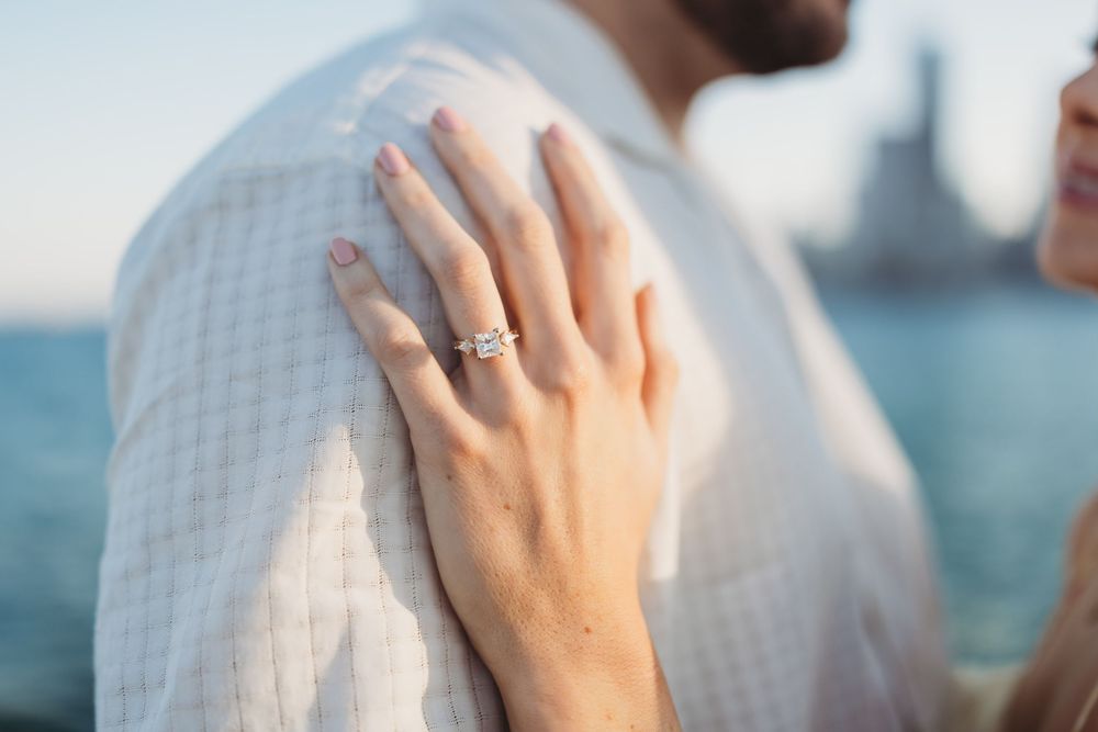 Close-up view of hands wearing a white knit sweater and engagement ring against an ocean backdrop.