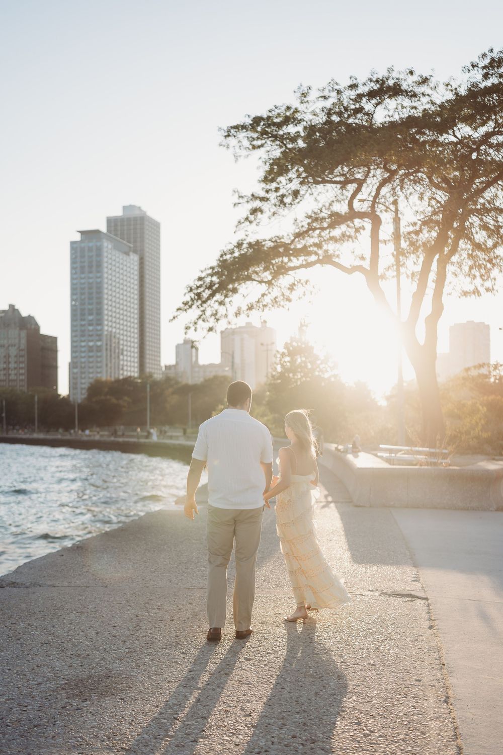 Silhouettes of a couple walking along waterfront path at golden hour with city buildings in background.