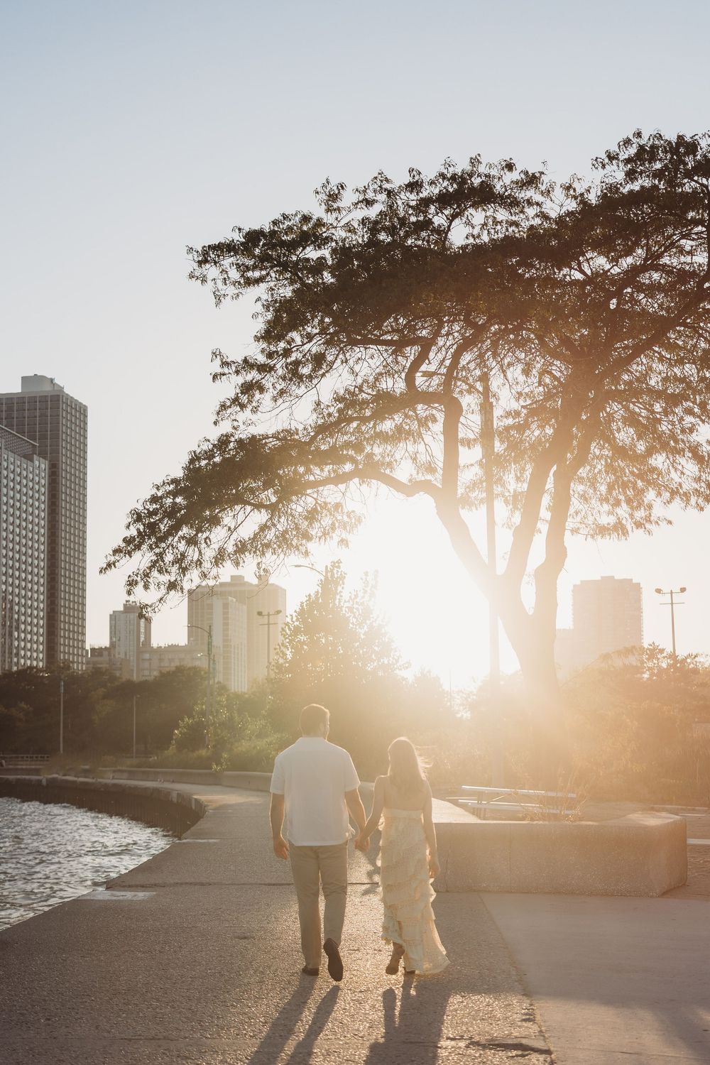 Two people walking along waterfront path at sunset with city skyline and large tree silhouetted in golden light.