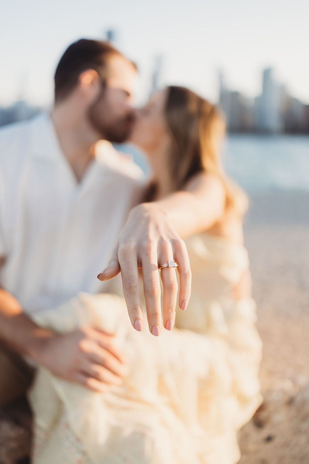 Close-up engagement ring shot with blurred couple and city skyline in background.