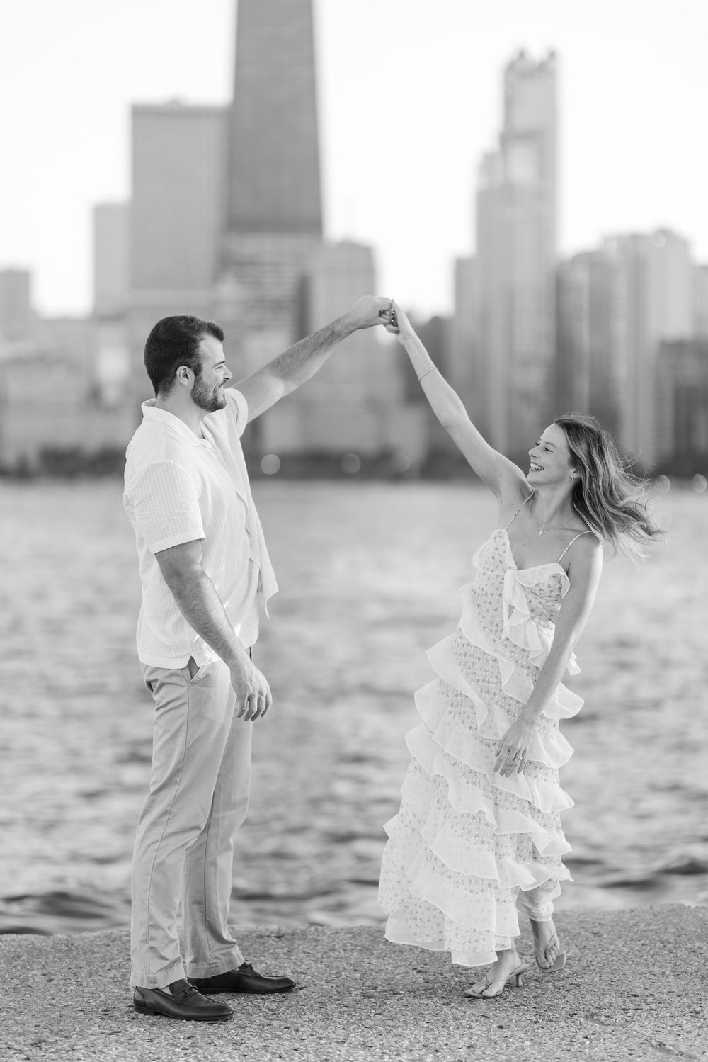 Black and white photo of a playful couple dancing on the shore with downtown Chicago's skyline visible.