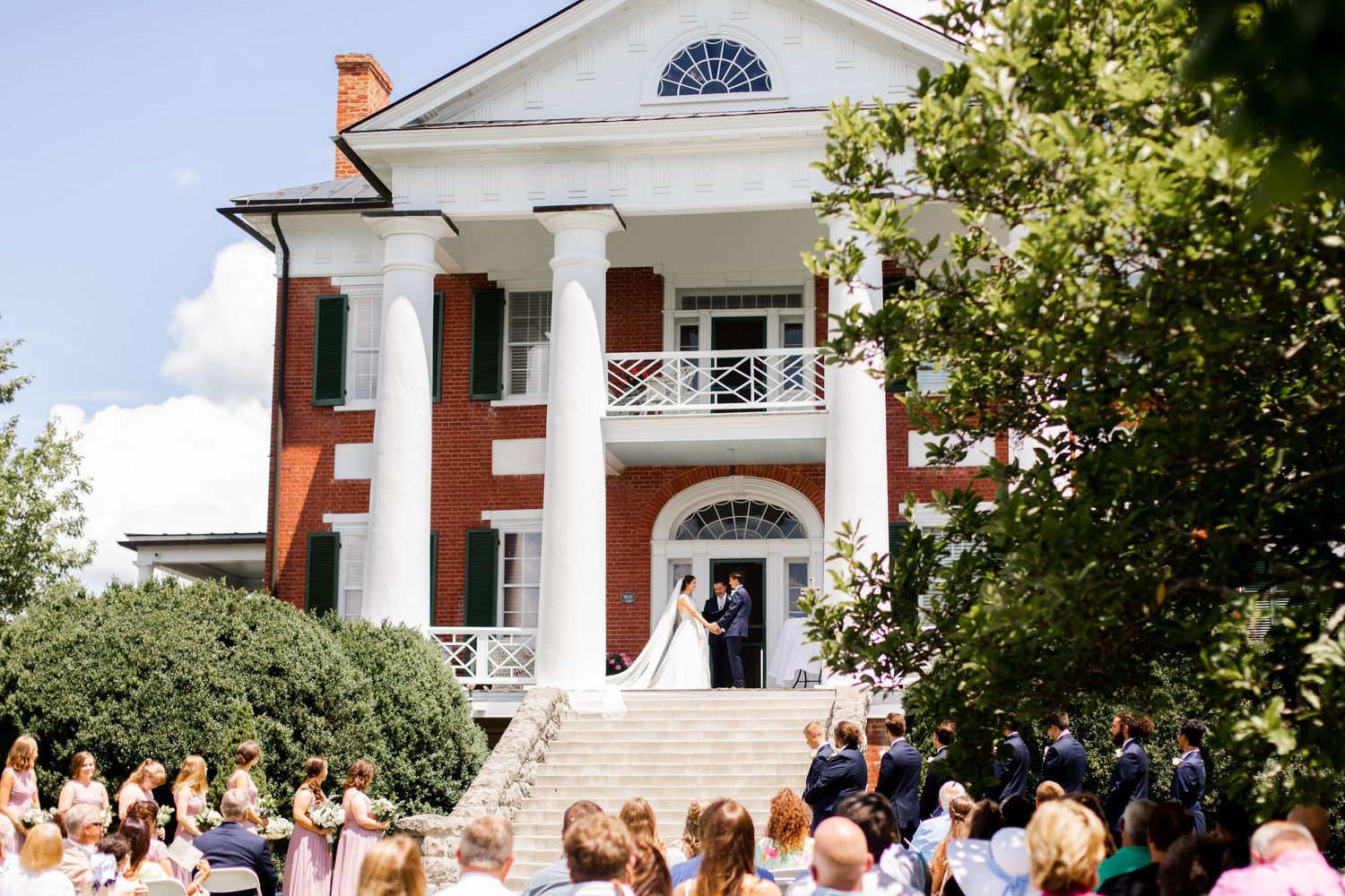 Outdoor wedding ceremony at a grand red brick colonial mansion with guests seated on the lawn.