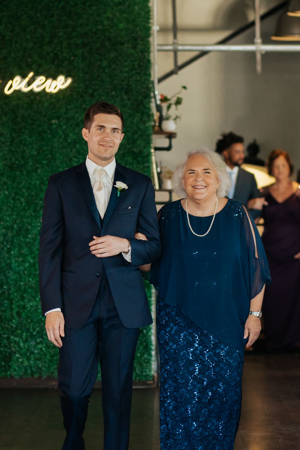 Formal portrait of a young man in navy suit and an older woman in blue lace dress against a green wall with neon sign.