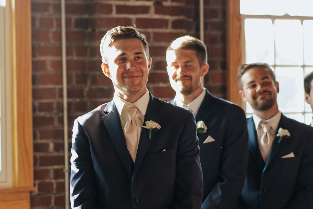 Groomsmen in dark suits pose together in front of a brick wall with sunlit windows during a wedding celebration.