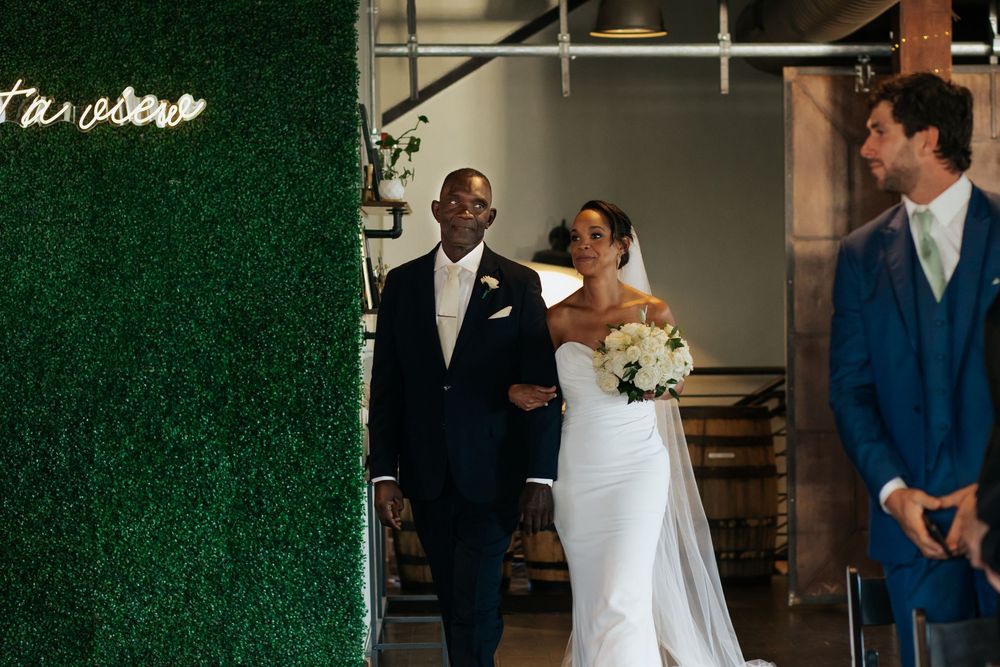 Wedding couple and groomsman share joyful moment in front of green hedge wall with neon signage.