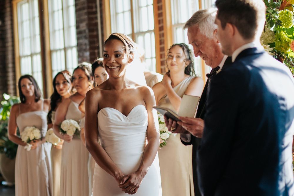 Wedding party stands together during ceremony in sunlit venue with large windows and beige bridesmaids dresses.
