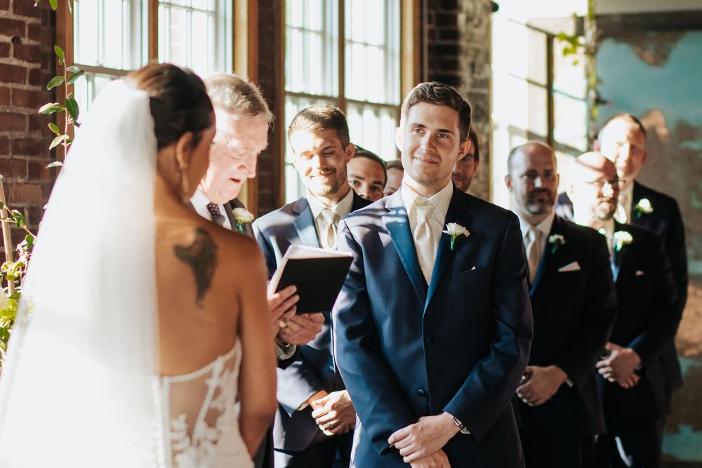 Wedding couple exchanges vows during indoor ceremony with groomsmen in navy suits looking on.