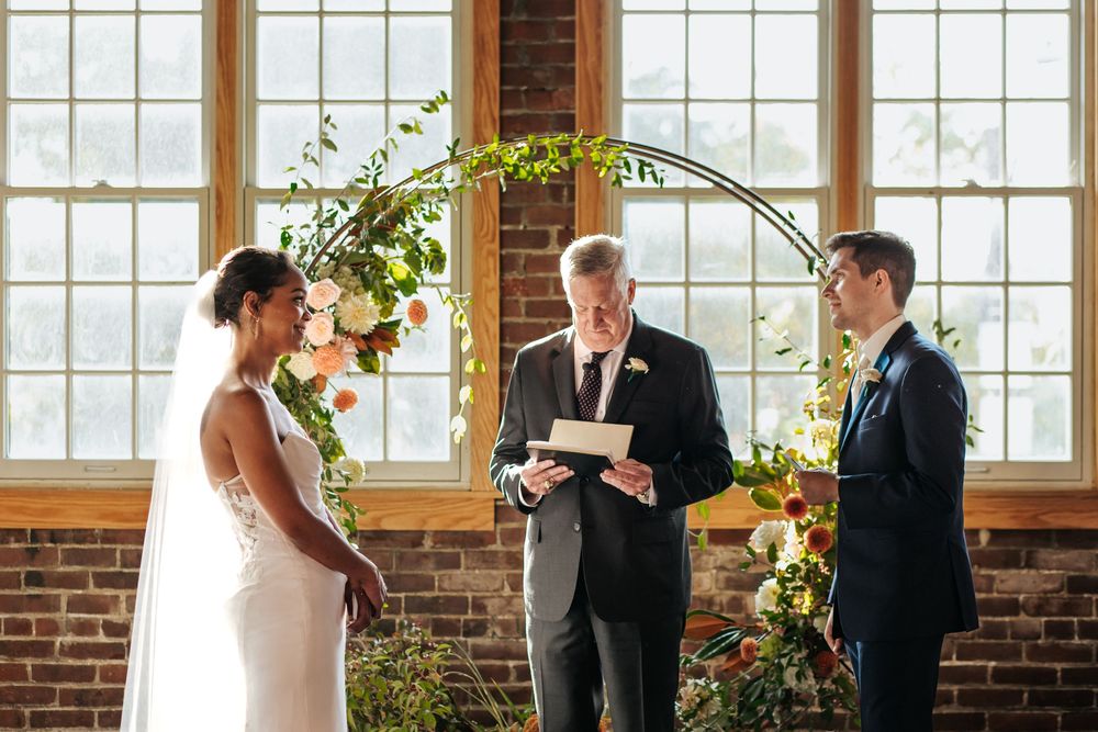 A wedding ceremony takes place in a rustic brick venue with large windows and natural lighting.