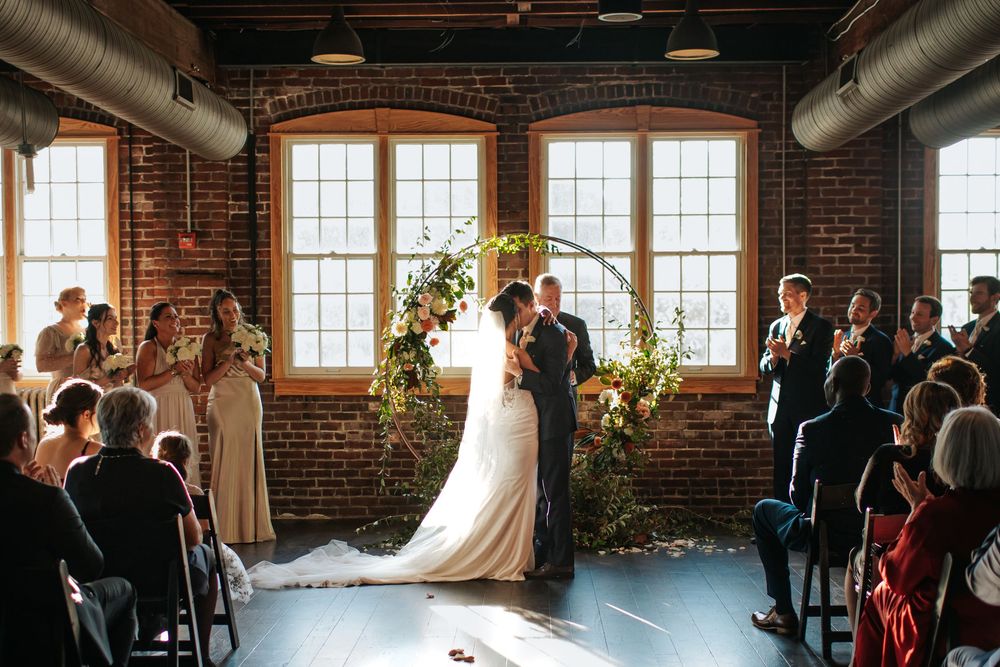 Wedding guests watch ceremony in a historic brick loft space with natural light streaming through tall windows.